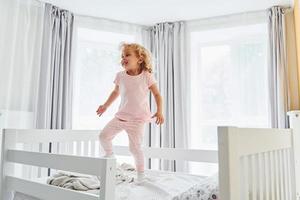saltando en la cama. una niña linda con ropa informal está en el interior de su casa durante el día foto