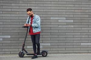 un joven apuesto con ropa informal de pie con un scooter eléctrico al aire libre durante el día soleado foto