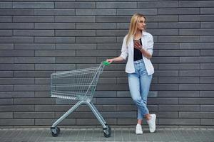 hermosa rubia con ropa informal con carrito de compras está al aire libre durante el día soleado foto