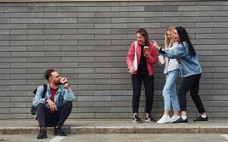 Three women standing and laughing at guy that sitting outdoors near building at daytime photo