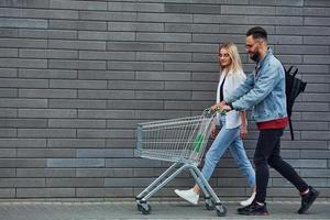 With shopping cart. Young stylish man with woman in casual clothes outdoors together. Conception of friendship or relationships photo