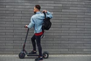 vista lateral de un joven apuesto con ropa informal que monta una moto eléctrica al aire libre durante el día soleado foto