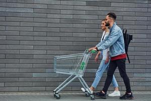 With shopping cart. Young stylish man with woman in casual clothes outdoors together. Conception of friendship or relationships photo
