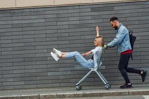 Having fun and riding shopping cart. Young stylish man with woman in casual clothes outdoors together. Conception of friendship or relationships photo