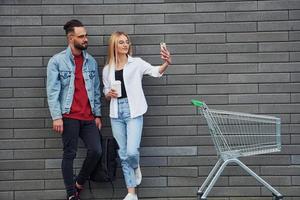 With shopping cart. Young stylish man with woman in casual clothes outdoors together. Conception of friendship or relationships photo