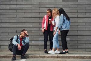 Three women standing and laughing at guy that sitting outdoors near building at daytime photo