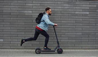 vista lateral de un joven apuesto con ropa informal que monta una moto eléctrica al aire libre durante el día soleado foto