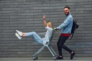 Having fun and riding shopping cart. Young stylish man with woman in casual clothes outdoors together. Conception of friendship or relationships photo