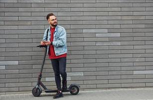 un joven apuesto con ropa informal de pie con un scooter eléctrico al aire libre durante el día soleado foto