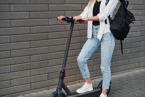 vista de cerca de una chica con ropa informal que está de pie con un scooter eléctrico al aire libre durante el día soleado foto