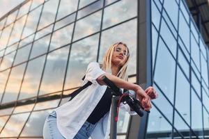 Against modern business building. Beautiful blonde in casual clothes riding electric schooter outdoors at sunny daytime photo