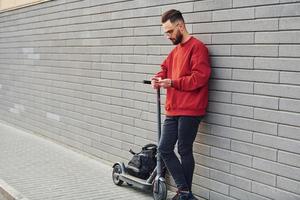 con bolso negro. un joven apuesto con ropa informal de pie con un scooter eléctrico al aire libre durante el día soleado foto