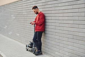 With black bag. Handsome young guy in casual clothes standing with electric schooter outdoors at sunny daytime photo