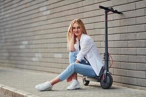 Beautiful blonde in casual clothes sitting on electric schooter outdoors at sunny daytime photo