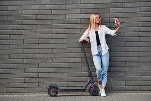 bella rubia con ropa informal parada con una escuela eléctrica al aire libre durante el día soleado con el teléfono en las manos foto