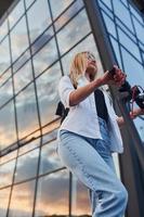 Against modern business building. Beautiful blonde in casual clothes riding electric schooter outdoors at sunny daytime photo