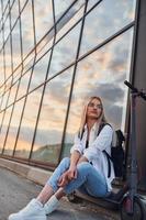 Against modern business building. Beautiful blonde in casual clothes riding electric schooter outdoors at sunny daytime photo