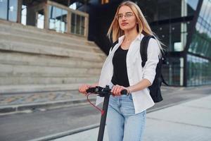 Near business building. Beautiful blonde in casual clothes riding electric schooter outdoors at sunny daytime photo