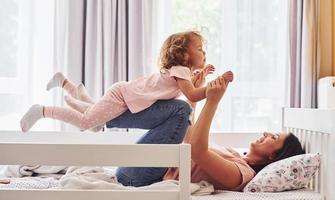 Playing with each other at weekend time. Young mother with her little daughter in casual clothes together indoors at home photo