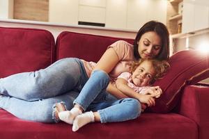 Lying down on red sofa. Young mother with her little daughter in casual clothes together indoors at home photo
