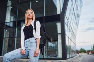 Beautiful blonde in casual clothes is outdoors at sunny daytime against business building photo