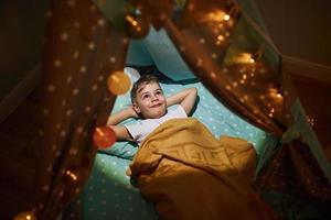 Young boy in casual clothes lying down in tent at evening time photo