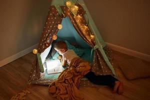 Reading book and using flashlight. Young boy in casual clothes lying down near tent at evening time photo