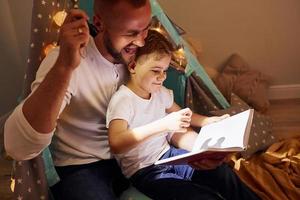 libro de lectura y uso de linterna. padre y su joven pasan tiempo juntos en el interior cerca de la tienda foto