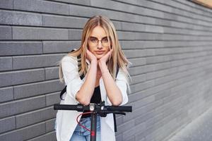 Against grey wall. Beautiful blonde in casual clothes riding electric schooter outdoors at sunny daytime photo