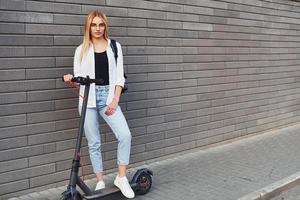 Against grey wall. Beautiful blonde in casual clothes riding electric schooter outdoors at sunny daytime photo