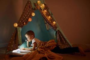 Reading book and using flashlight. Young boy in casual clothes lying down near tent at evening time photo