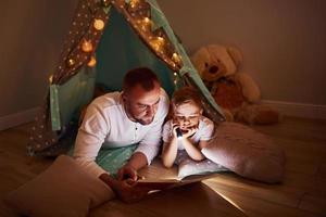 libro de lectura y uso de linterna. padre y su joven pasan tiempo juntos en el interior cerca de la tienda foto