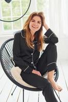 Cheerful young woman in black pajamas sitting on chair indoors at daytime photo