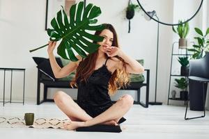 Young woman in pajamas sitting on the floor indoors at daytime and holding big green leaf photo