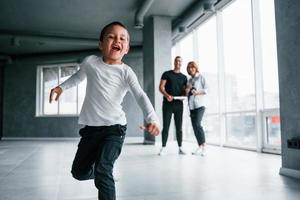 Woman in formal clothes and with camera in hand standing inside of empty room with man and little boy that running and having fun photo