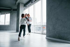 Woman in formal clothes and with camera in hand standing inside of empty room with man and little boy that running and having fun photo