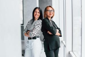 Two business women standing together indoors in the office near big window photo