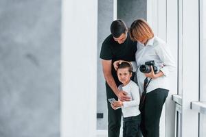 Woman in formal clothes and with camera in hand standing inside of empty room with man and little boy photo