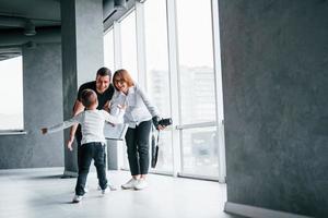 Woman in formal clothes and with camera in hand standing inside of empty room with man and little boy that running and having fun photo