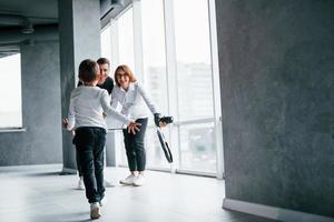 Woman in formal clothes and with camera in hand standing inside of empty room with man and little boy that running and having fun photo