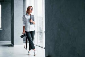 Woman in formal clothes and with camera in hand standing inside of empty room photo