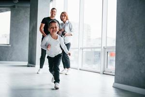 Woman in formal clothes and with camera in hand standing inside of empty room with man and little boy that running and having fun photo