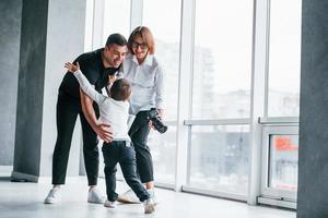 Woman in formal clothes and with camera in hand standing inside of empty room with man and little boy that running and having fun photo