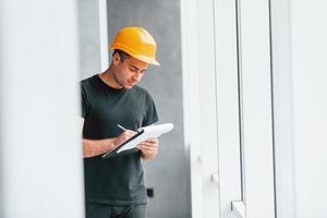 Male worker or engineer in yellow hard hat standing indoors with notepad photo