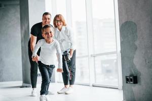 Woman in formal clothes and with camera in hand standing inside of empty room with man and little boy that running and having fun photo