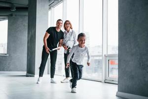 Woman in formal clothes and with camera in hand standing inside of empty room with man and little boy that running and having fun photo