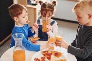 tres niños sentados en el interior junto a la mesa y comiendo pizza con jugo de naranja juntos foto
