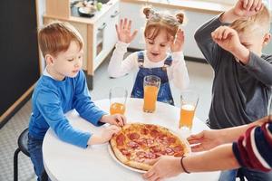 Three children sitting indoors by the table and eating pizza with orange juice together photo