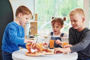 Three children sitting indoors by the table and eating pizza with orange juice together photo