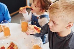 tres niños sentados en el interior junto a la mesa y comiendo pizza con jugo de naranja juntos foto